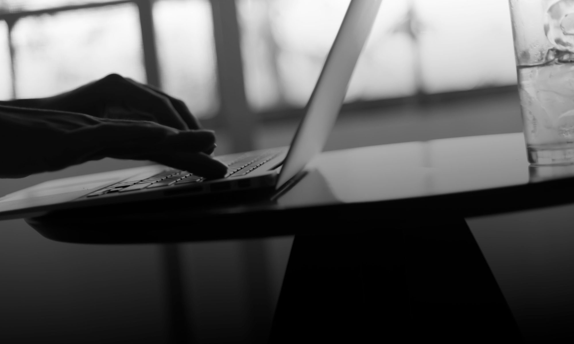 Close-up of a person's hands typing on a laptop keyboard with a glass of water on the table, and a blurred background. Black and white photo.